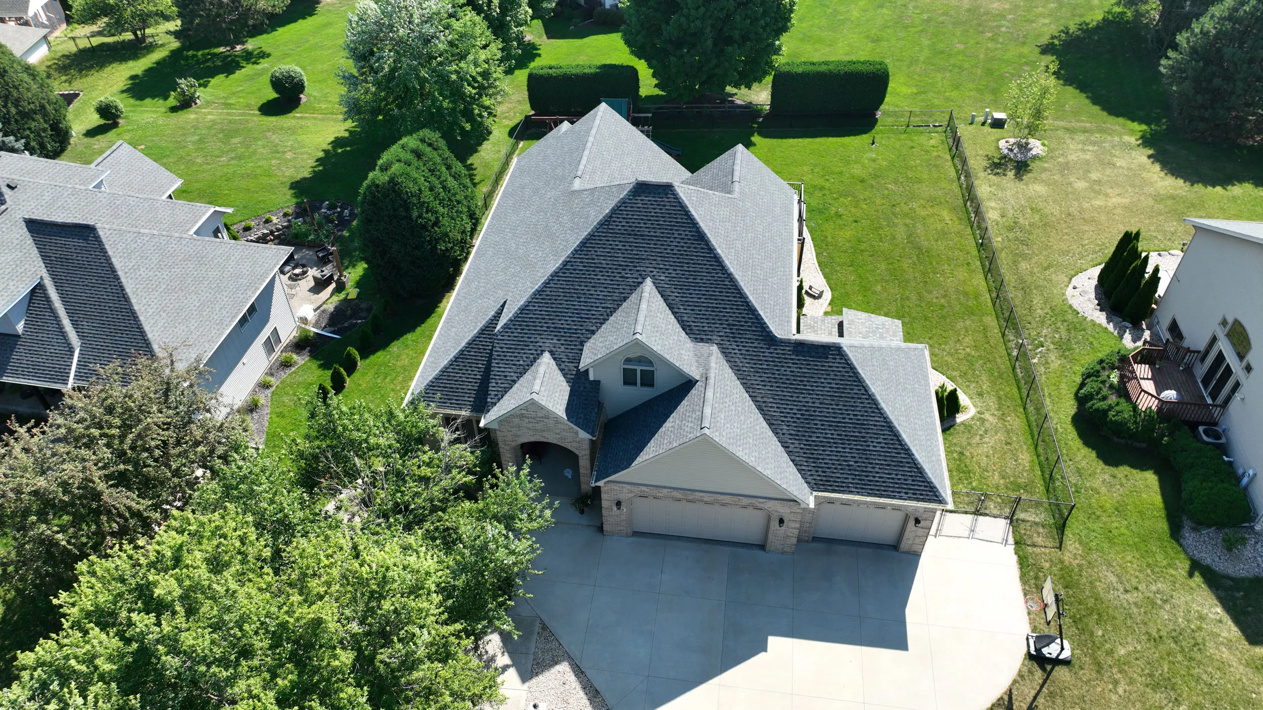 Aerial view of a large suburban house with a gray roof, expansive driveway, and green landscaped yard.